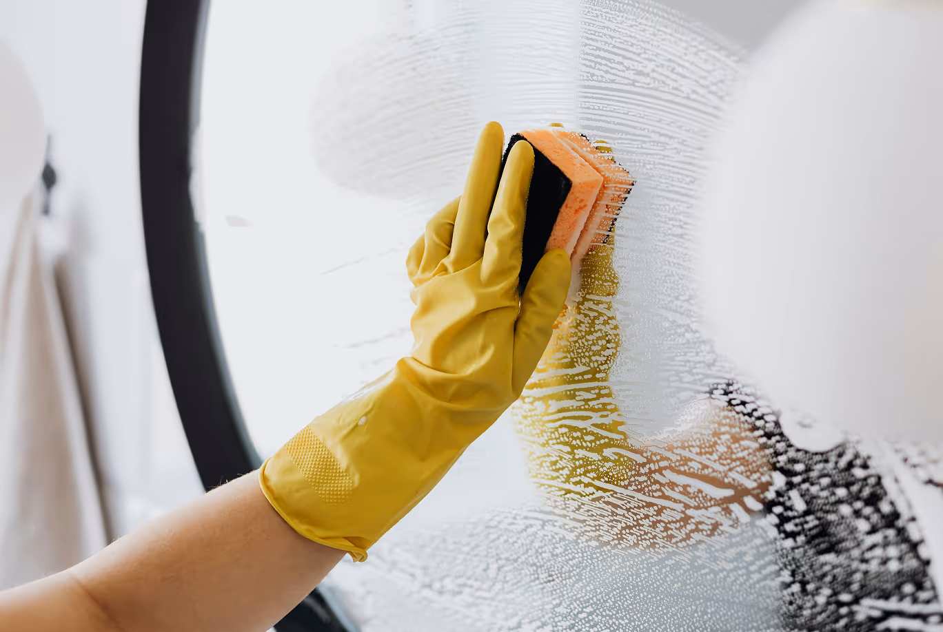Person wearing yellow gloves pouring liquid detergent onto a black and orange cleaning sponge.