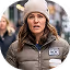 Smiling woman wearing a beige beanie and warm jacket, holding a microphone at an outdoor event with a crowd in the background.